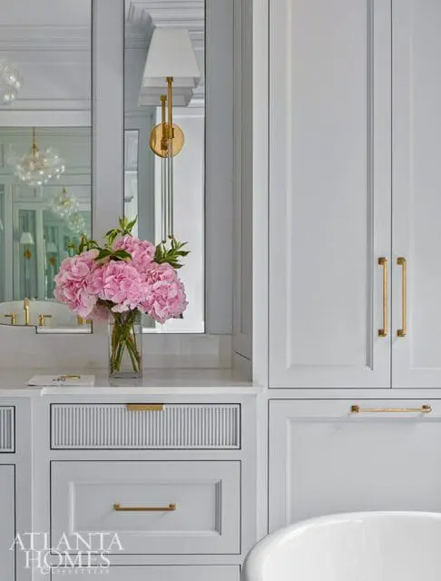 Light gray bathroom vanity with fluted and paneled drawers, brass hardware, and a vase of pink hydrangeas on a marble countertop.