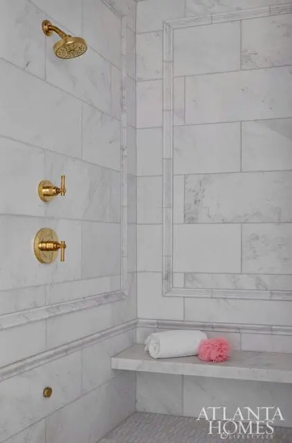 Marble tiled shower with gold fixtures, including a shower head and two handles. A built-in marble bench holds a white towel and a pink loofah.