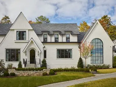White stucco house with dark-framed windows, gabled rooflines, and a large arched window. Landscaped lawn and stone retaining wall in front.