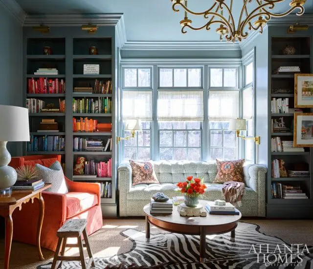 Light blue living room with floor-to-ceiling bookshelves, tufted sofa, orange armchair, zebra rug, and oval coffee table with floral arrangement