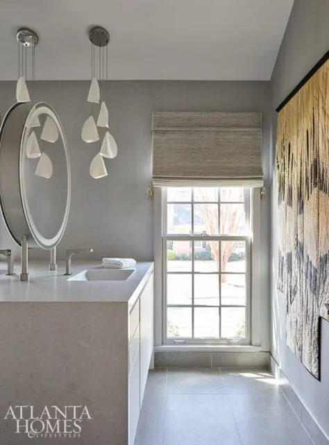 Gray bathroom with a floating vanity, oval mirror with integrated lighting, modern pendant lights, and a large window with a Roman shade.