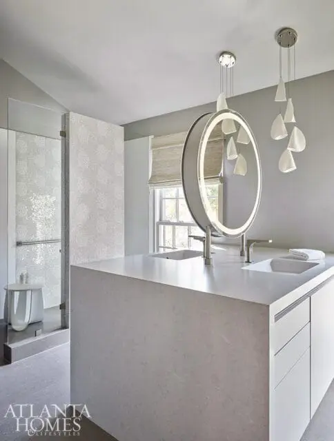 Modern bathroom with a large gray stone vanity featuring two sinks, a circular backlit mirror, and contemporary pendant lighting. A glass-enclosed shower is visible to the left.