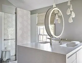 Modern bathroom with a large circular vanity mirror with integrated lighting, a light gray countertop with two sinks, and a glass-enclosed shower with textured walls.