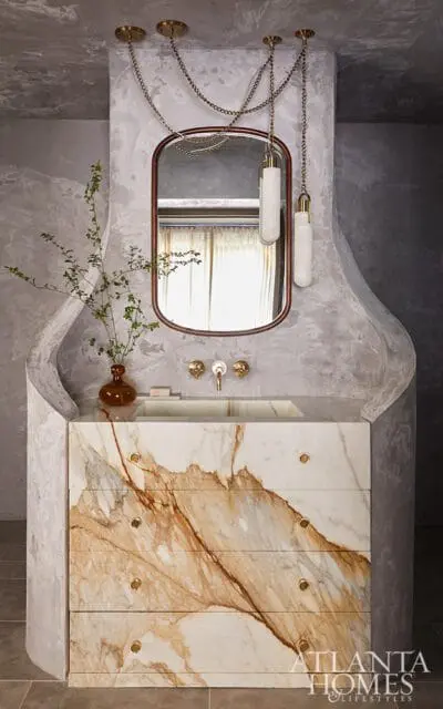 Bathroom vanity with a marble countertop and drawers featuring dramatic brown veining, a rectangular sink, and brass fixtures. Above, a mirror is framed by a dark wood border, and two pendant lights hang from chains.