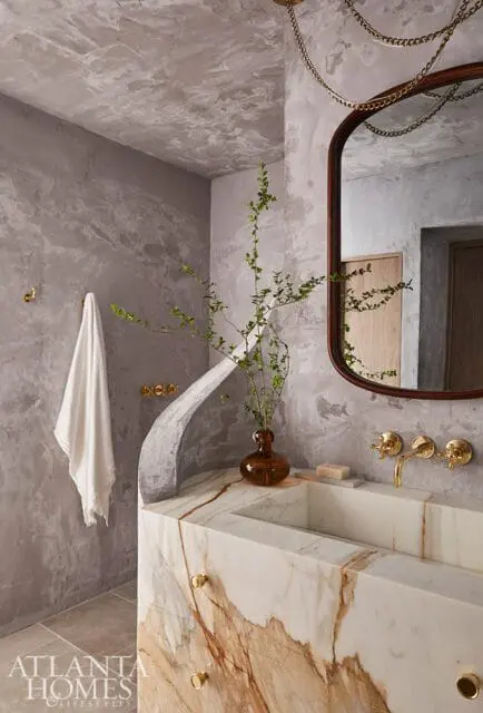 Modern bathroom with textured gray walls, a marble vanity with brass fixtures, and a rounded mirror with a wooden frame.