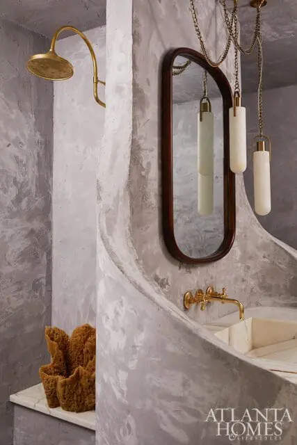 Bathroom with textured gray walls, brass showerhead, oval mirror with dark wood frame, and a curved vanity with a brass faucet.