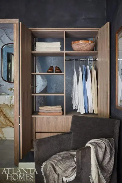 Open wood closet with folded towels, shoes, a basket, and hanging clothes. A dark armchair with a blanket is in the foreground.
