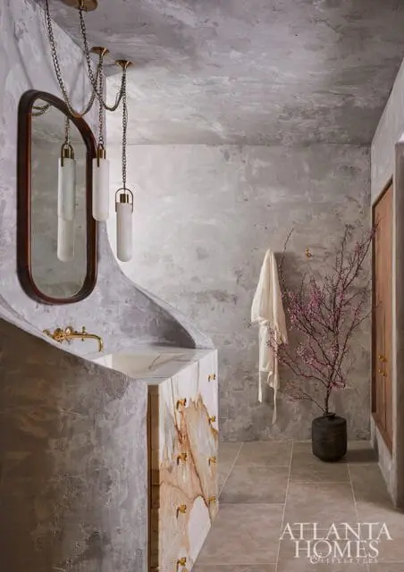 Bathroom with textured gray walls, a marble vanity with brass hardware, an oval mirror, and a flowering branch in a vase.