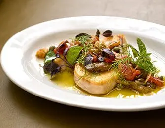 Seared scallops topped with chopped tomatoes, capers, and fresh herbs, served in a white oval dish with olive oil