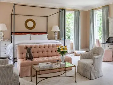 Bedroom with a black canopy bed, pink tufted sofa, two armchairs, a vintage secretary desk, and a French bulldog sitting on the sofa.