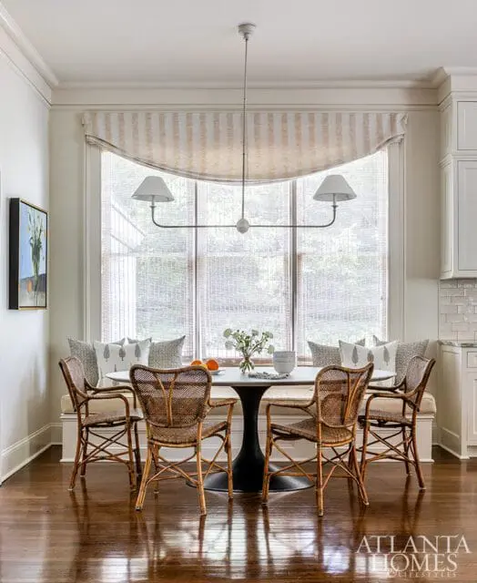Round dining table with woven chairs and banquette seating in front of a large window with a striped valance and woven shades.