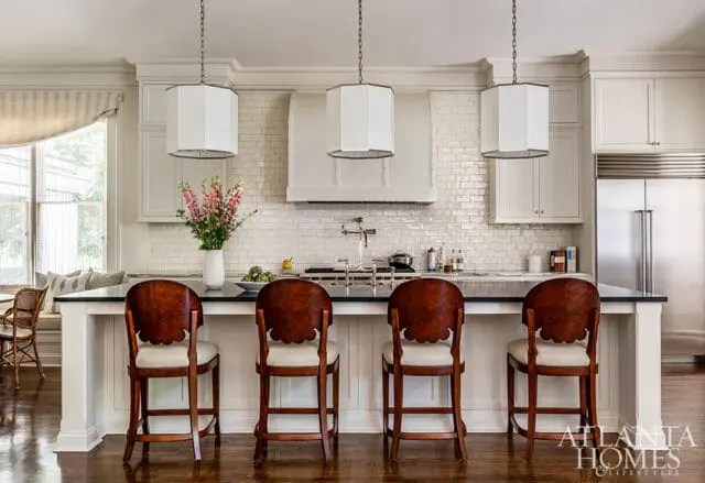 White subway tile backsplash in a kitchen with a large range hood, white cabinetry, and a dark countertop with four wooden bar stools.