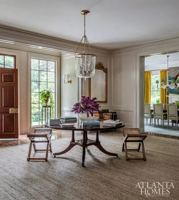 Foyer with a dark wood door, large windows, a round pedestal table with books and purple flowers, two wicker stools, and a jute rug. A dining room is visible through an open doorway.