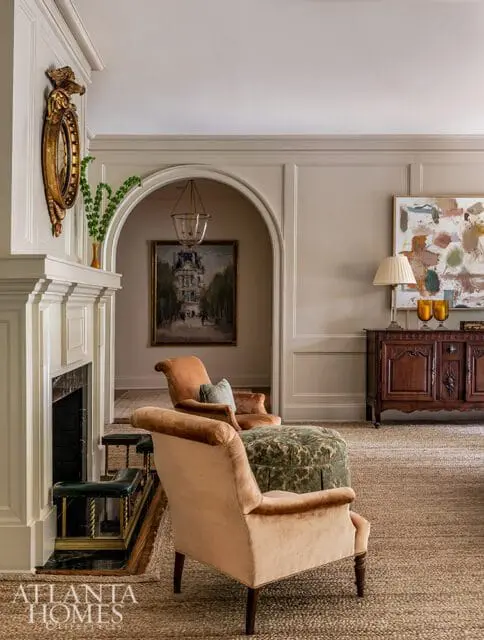 Living room with a fireplace, two velvet armchairs in peach and rust tones, a green patterned ottoman, and a dark wood cabinet with abstract art above.