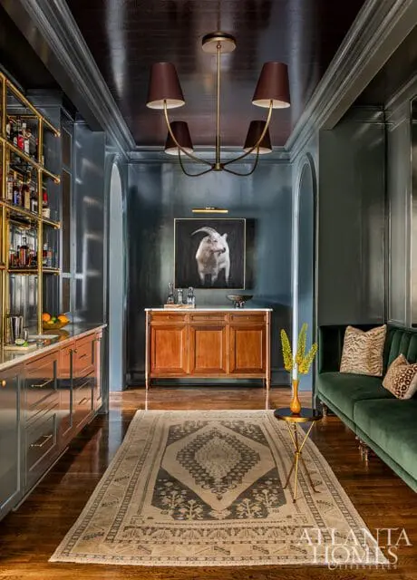 Dark blue walls with a gold-framed painting of a goat above a wooden credenza, flanked by a bar area and a green velvet sofa.