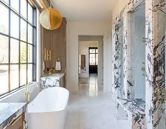 Luxury bathroom with a freestanding white tub, marble walls and countertops, brass fixtures, and a large window with a gold pendant light.
