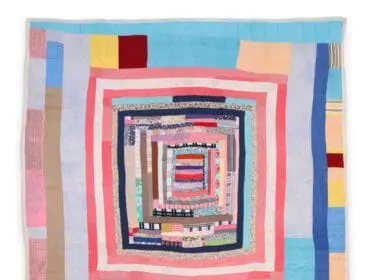 A handmade quilt with a log cabin pattern, featuring concentric squares of fabric scraps in various colors and patterns, including pink, blue, yellow, red, and brown.