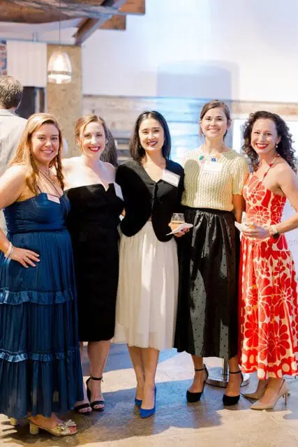 Five women posing for a photo at an event. From left to right: a woman in a blue tiered dress, a woman in a black strapless dress, a woman in a black jacket and white skirt, a woman in a yellow textured top and black skirt, and a woman in a red floral dress.