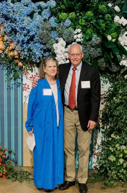 A woman in a blue coat and a man in a suit and tie stand in front of a floral backdrop.