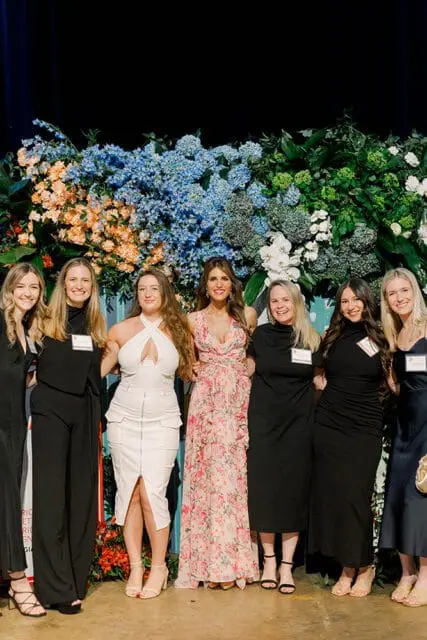 Seven women stand in front of a floral backdrop featuring blue hydrangeas, orange flowers, and greenery. The woman in the center wears a pink floral dress, while others wear black or white attire.