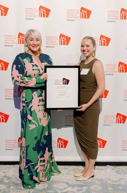 Two women stand in front of a backdrop with the American Society of Interior Designers Georgia logo. The woman on the left wears a green, pink, and navy floral dress and holds a framed award. The woman on the right wears a brown sleeveless dress and has a name tag.