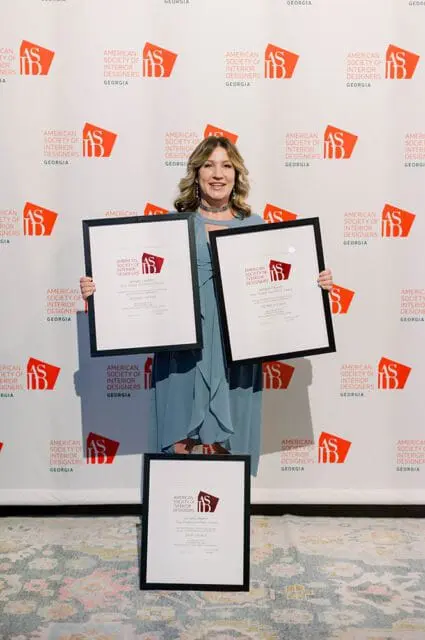 Woman in blue dress holding three framed awards from the American Society of Interior Designers Georgia Chapter, standing in front of a backdrop with the ASID Georgia logo.