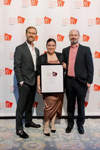 Three people stand in front of a backdrop with the American Society of Interior Designers Georgia logo. The person in the middle holds a framed award.