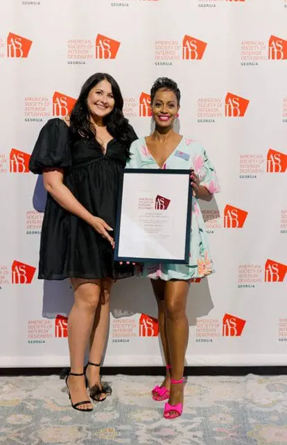 Two women stand in front of a backdrop with the American Society of Interior Designers Georgia logo. The woman on the left wears a black dress and the woman on the right wears a light blue dress with floral print and holds a framed award.