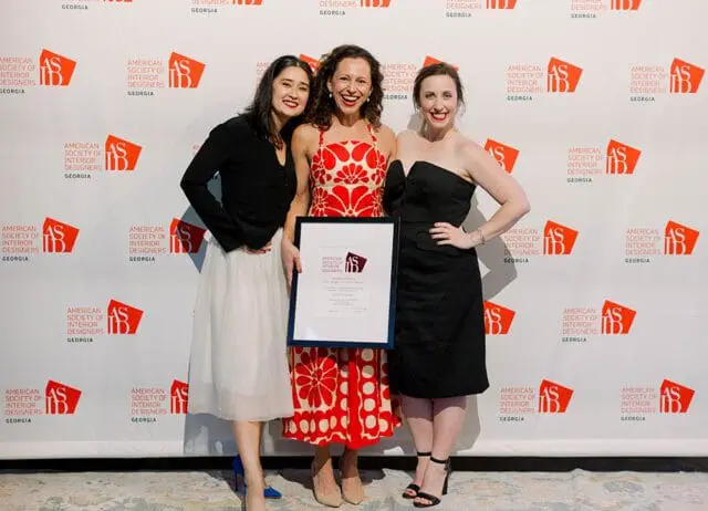Three women pose for a photo in front of a backdrop with the American Society of Interior Designers Georgia logo repeated. The woman in the center holds a framed award.
