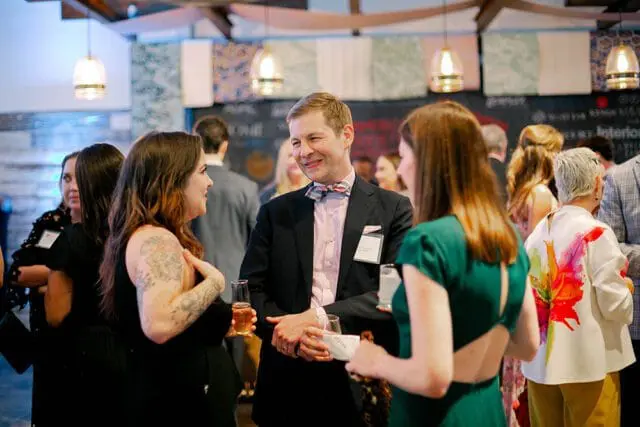 People mingling at an event, with a man in a suit and bow tie speaking to two women. The background features decorative wall hangings and pendant lights.