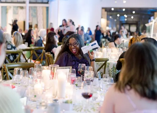 Woman in glasses smiles and holds up a card at a formal dinner event with candles and wine glasses on the table.