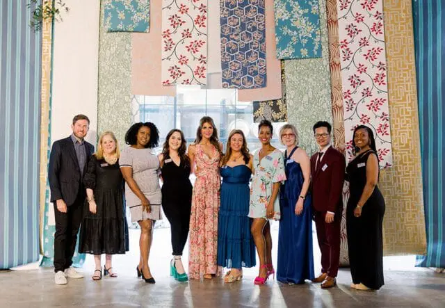 A group of nine people, including six women and three men, stand in front of a wall displaying various patterned wallpapers.