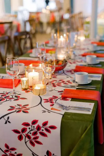 Dining table set with white plates, wine glasses, orange napkins, and floral patterned runner with red flowers and black vines. Candles in glass holders provide ambient light.