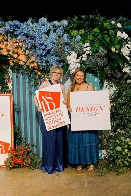 Two women stand in front of a floral display holding signs. The woman on the left holds a sign that reads "ASID American Society of Interior Designers." The woman on the right holds a sign that reads "The 13th Annual DESIGN Excellence Awards."