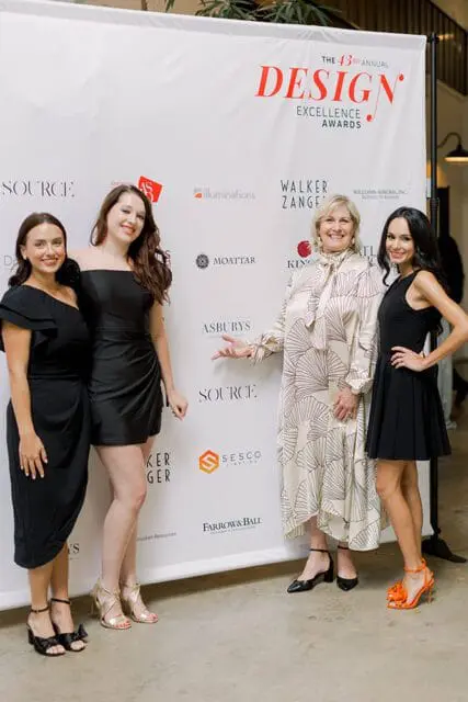 Four women pose in front of a white backdrop with "The 43rd Annual Design Excellence Awards" logo and sponsor names. The women wear black and patterned dresses.