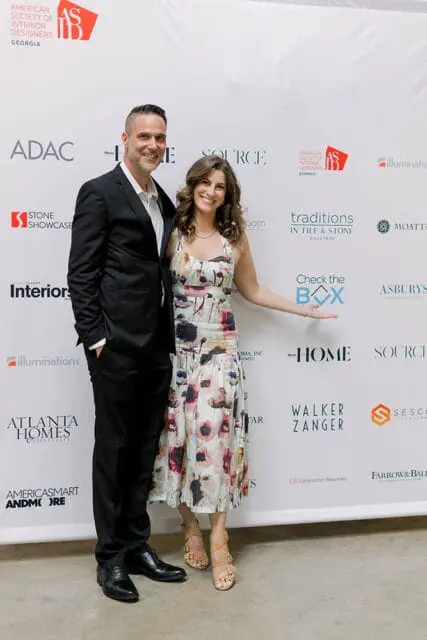 Man in a black suit and woman in a floral dress pose in front of a white backdrop with sponsor logos including ADAC, Stone Showcase, Atlanta Homes & Lifestyles, and Traditions in Tile & Stone.
