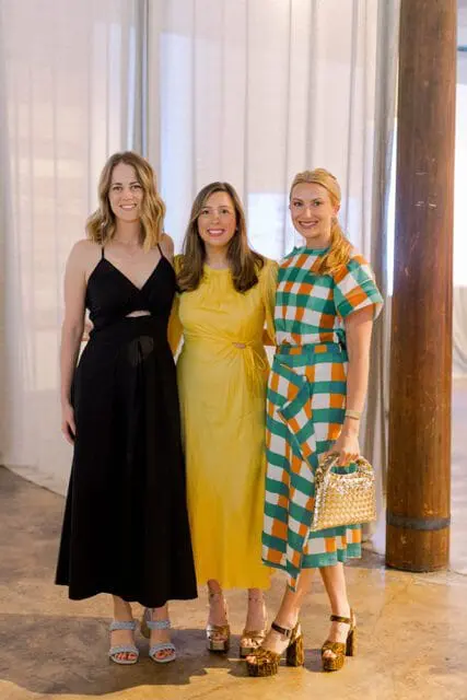 Three women standing together, the woman on the left wears a black dress, the middle woman wears a yellow dress, and the woman on the right wears a green and orange checkered dress and holds a gold purse.
