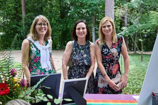 Three women stand near a computer display with floral arrangements.
