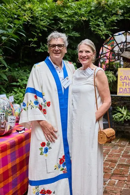 Man wearing a white robe with blue trim and floral embroidery, and a woman in a white dress, posing outdoors with lush greenery in the background.
