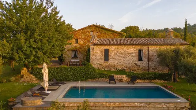 Stone villa with ivy-covered walls and a tiled roof, featuring a swimming pool with lounge chairs and umbrellas, surrounded by lush greenery and trees.