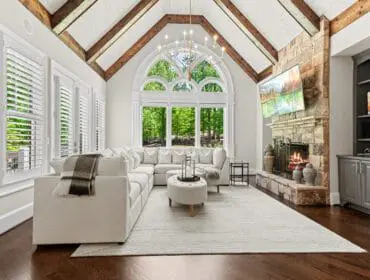 Living room with vaulted wood-beamed ceiling, large arched window overlooking trees, sectional sofa, stone fireplace with a television above, and dark hardwood floors.