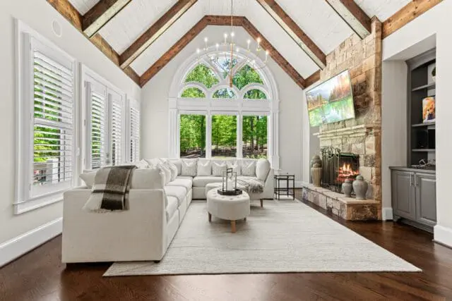 Living room with vaulted wood-beamed ceiling, large arched window overlooking trees, sectional sofa, stone fireplace with a television above, and dark hardwood floors.