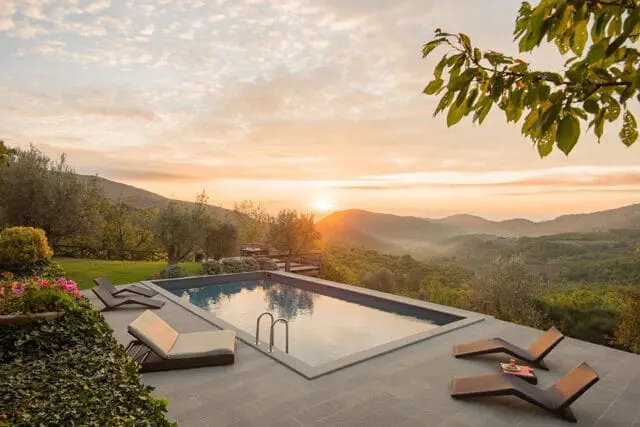 Rectangular swimming pool on a stone patio overlooking rolling hills at sunset. Two lounge chairs are placed on the patio.