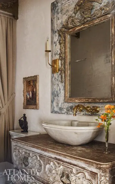 Bathroom vanity with carved wood base, marble vessel sink, brass faucet, ornate distressed mirror, and a small painting on the wall.