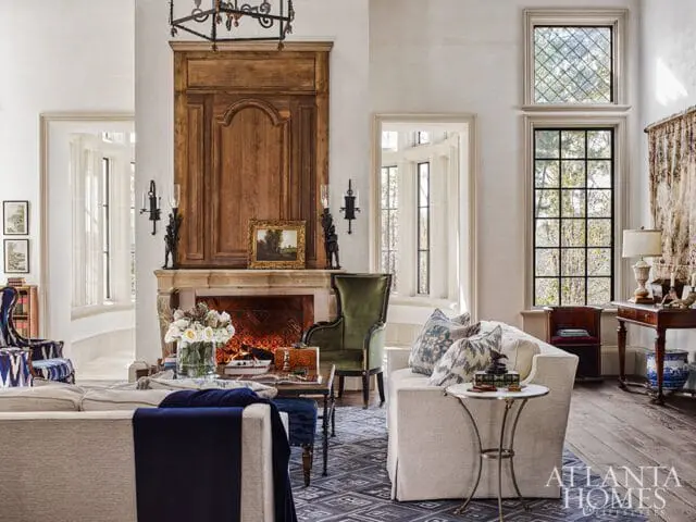 Living room with a large wooden fireplace, plush sofas, green velvet armchair, and tall windows overlooking trees.