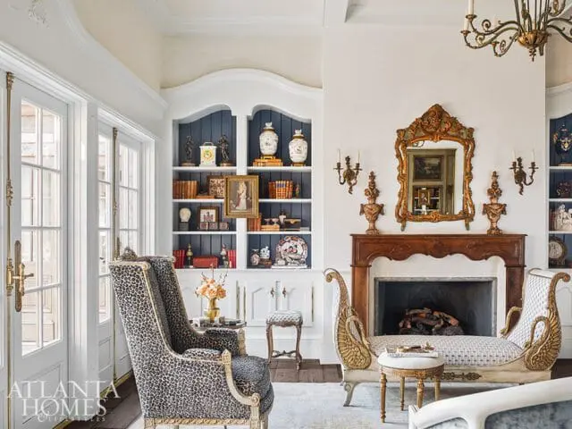 Living room with a leopard print armchair, ornate fireplace with a gilded mirror, built-in bookshelves, and French doors.