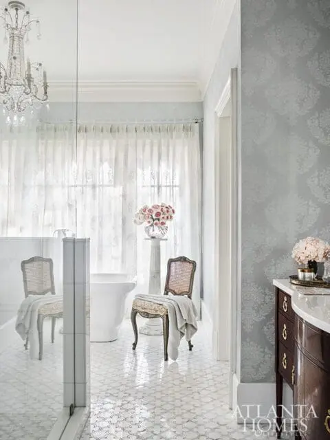 Elegant bathroom with a freestanding tub, ornate chandelier, patterned wallpaper, and a marble mosaic floor. Two cane-backed chairs with draped towels flank the tub.