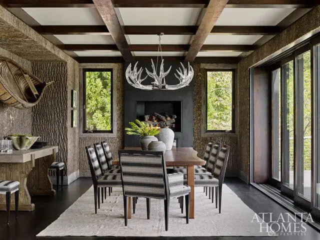 Rustic dining room with wood-beamed ceiling, bark-textured walls, a live-edge dining table with striped chairs, and a fireplace with an antler chandelier.