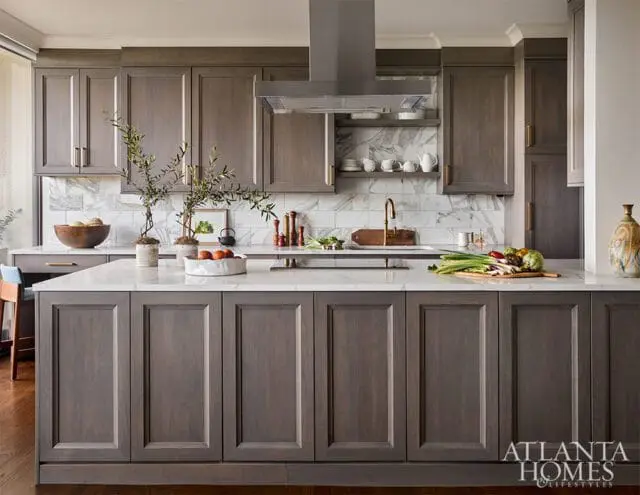 Gray wood kitchen cabinets with white marble backsplash and countertops. A large island features a sink and brass faucet, with fresh produce on display.