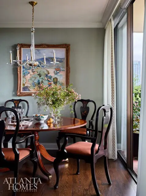 Dining room with a round wooden table, four dark wood chairs with coral cushions, a crystal chandelier, and a landscape painting on a muted green wall.